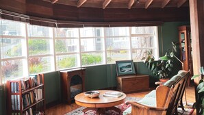 Een gezellige kamer met een houten plafond, een groot raam, een boekenplank, een ronde tafel en een comfortabele fauteuil.