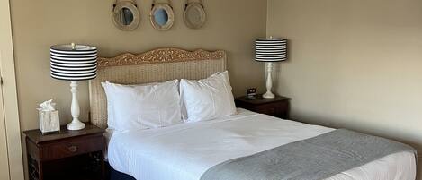 Una cama bien hecha con sábanas blancas y un plaid gris, flanqueada por dos mesitas de noche con lámparas, un tocador de madera y un espejo montado en la pared.