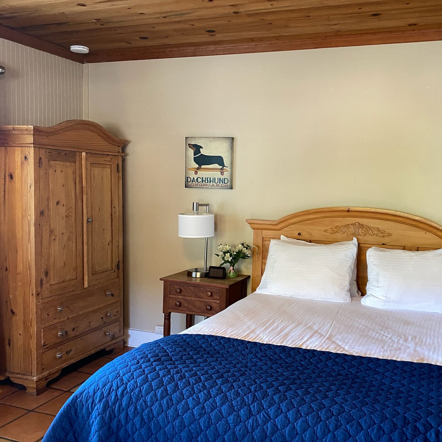 A bedroom with a wooden headboard, a bed with white linens, a blue quilted bedspread, a nightstand with a lamp and flowers, and a wooden wardrobe.