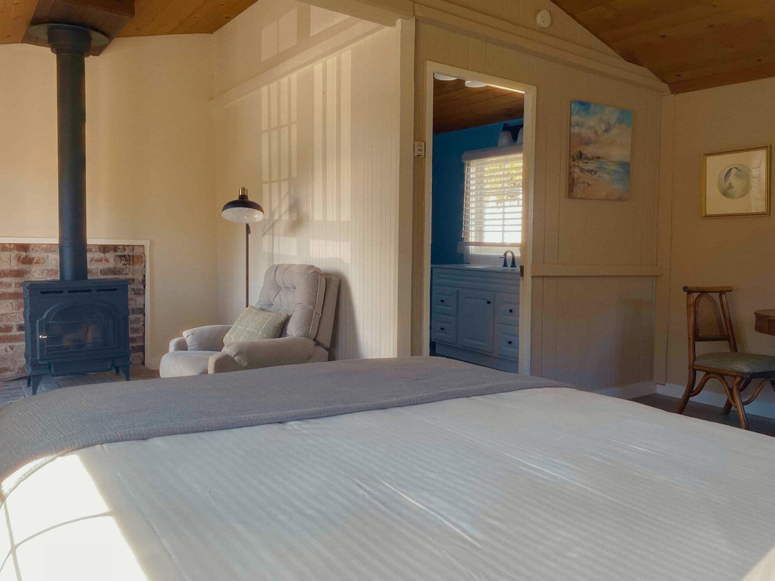 A bedroom with a bed, a chair, a lamp, a fireplace, and a view into another room.