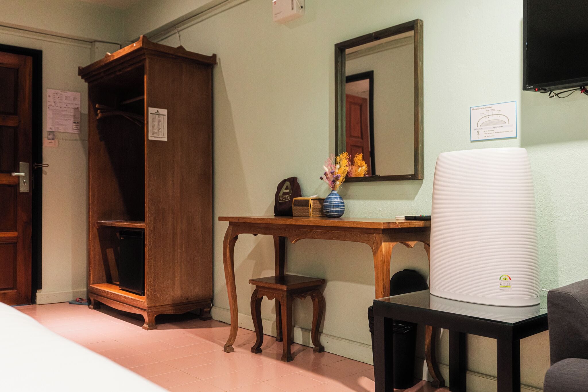 A hotel room with a wooden wardrobe, a small wooden table with a vase of flowers, a white air purifier, and a mirror on the wall.