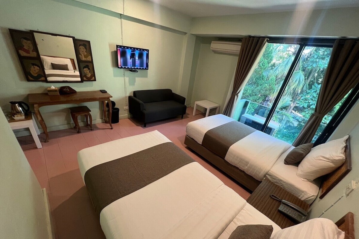 A hotel room with two beds, a desk, a couch, a TV, and a window with a view of greenery.