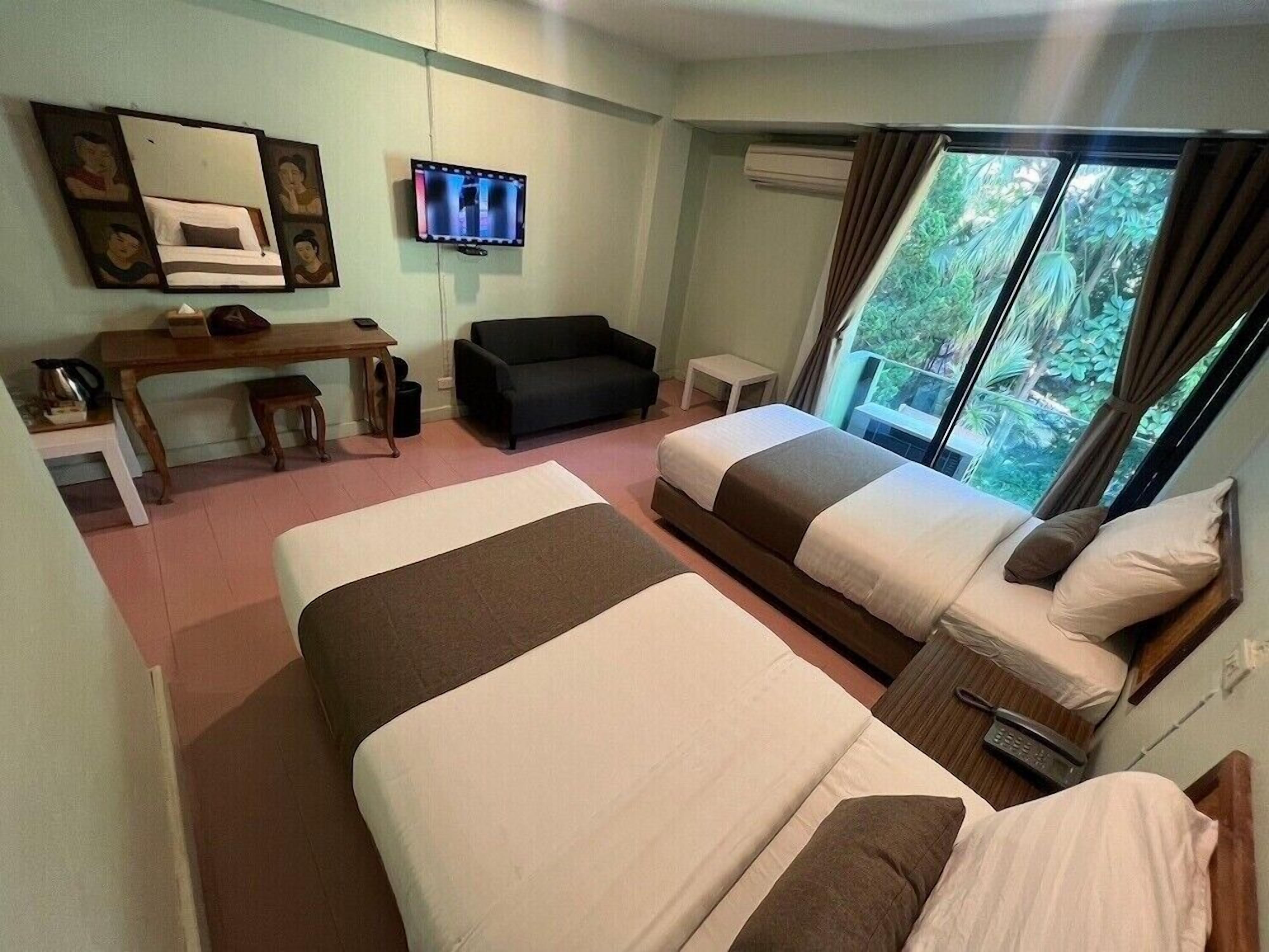 A hotel room with two beds, a desk, a couch, a TV, and a window with a view of greenery.