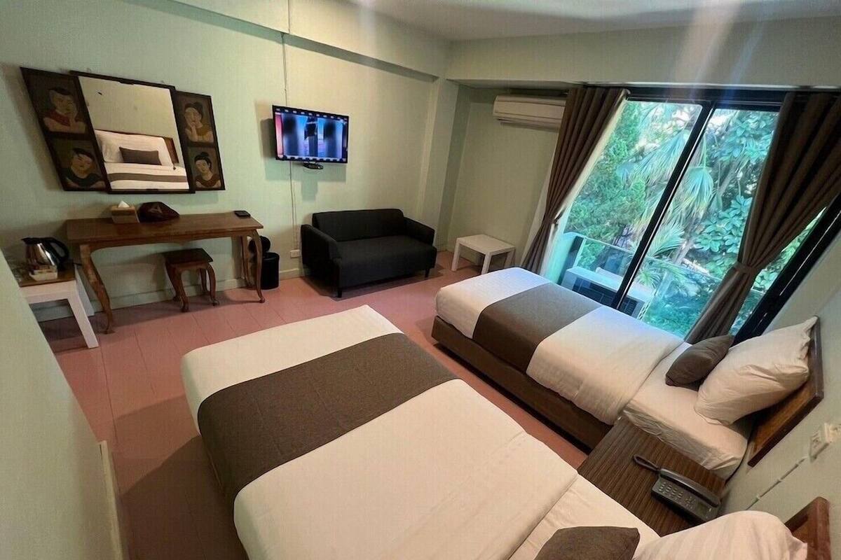 A hotel room with two beds, a desk, a couch, a TV, and a window with a view of greenery.