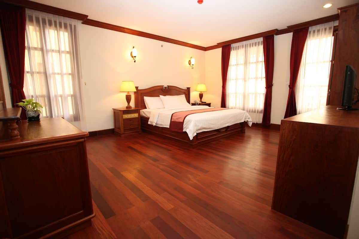 Ein Hotelzimmer mit einem hölzernen Bett, zwei Nachttischen mit Lampen, einer Holzkommode und einem großen Fenster mit Vorhängen.