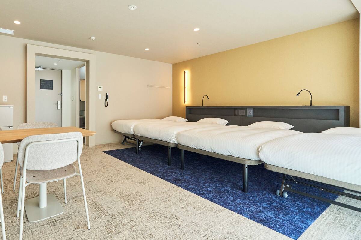 A hotel room with a desk, two chairs, a bed with two pillows, a bedside table, and a wall-mounted lamp.