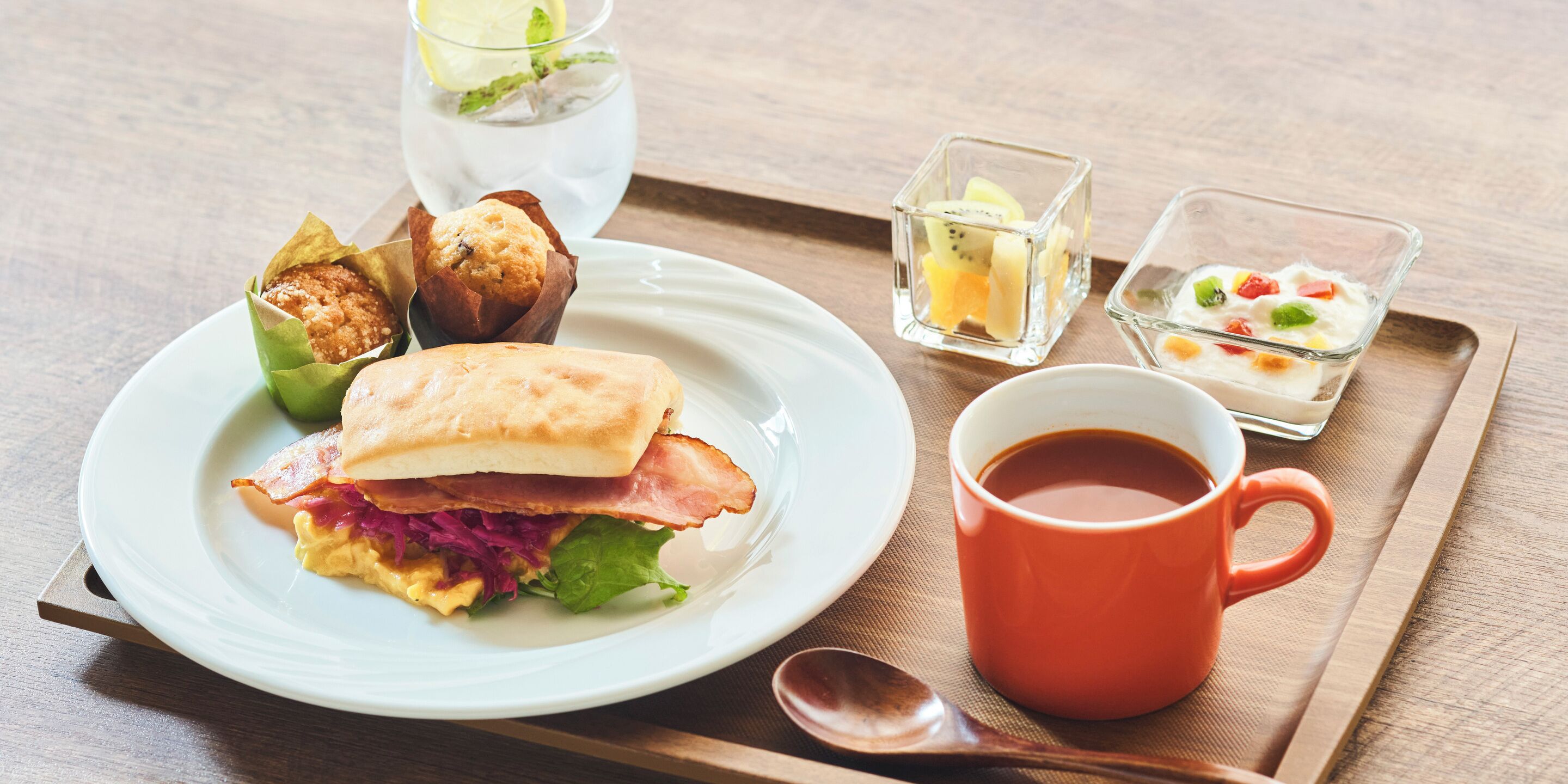 Bandeja de desayuno con un sándwich, un muffin, fruta y una taza de café.