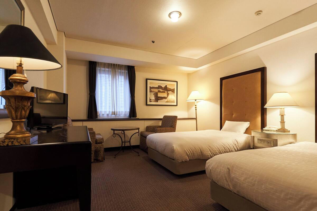 A hotel room with two beds, a desk, a chair, a TV, and a window with curtains.