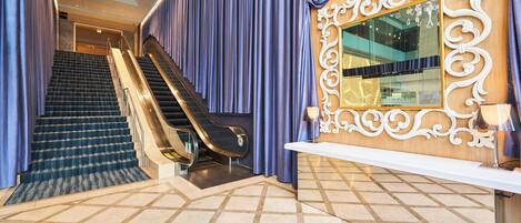 Un vestíbulo de hotel con una escalera monumental, una recepción y un espejo decorativo.