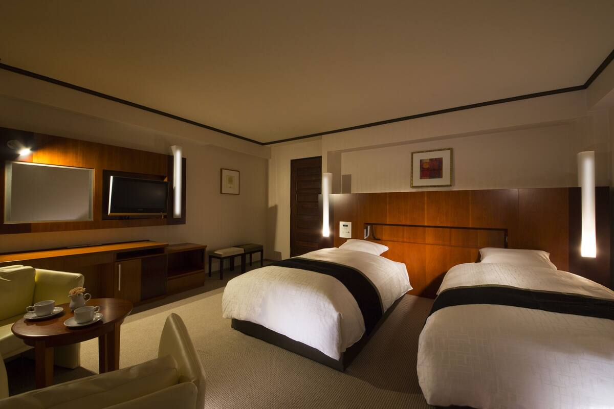 A hotel room with two beds, a desk with a chair, a television, and a small table with cups.