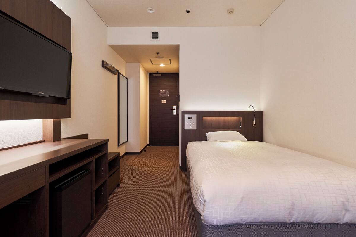A hotel room with a bed, a desk with a flat-screen TV, a door, and a wall-mounted light.