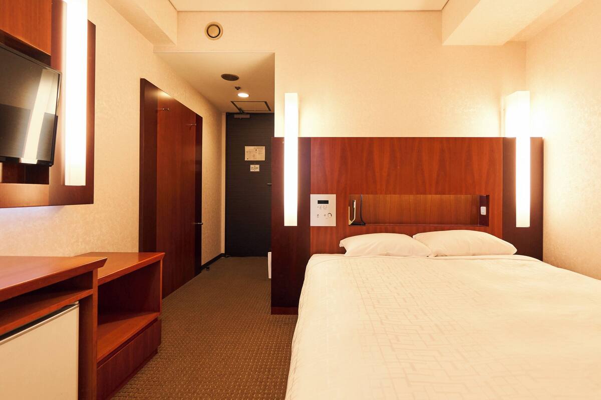 A hotel room with a bed, a TV mounted on the wall, a desk, and a wooden headboard.