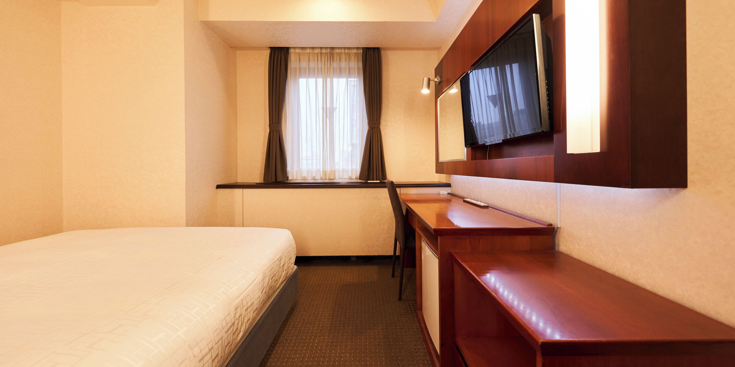 Habitación de hotel con cama, escritorio, silla, televisor montado en la pared y ventana con cortinas.