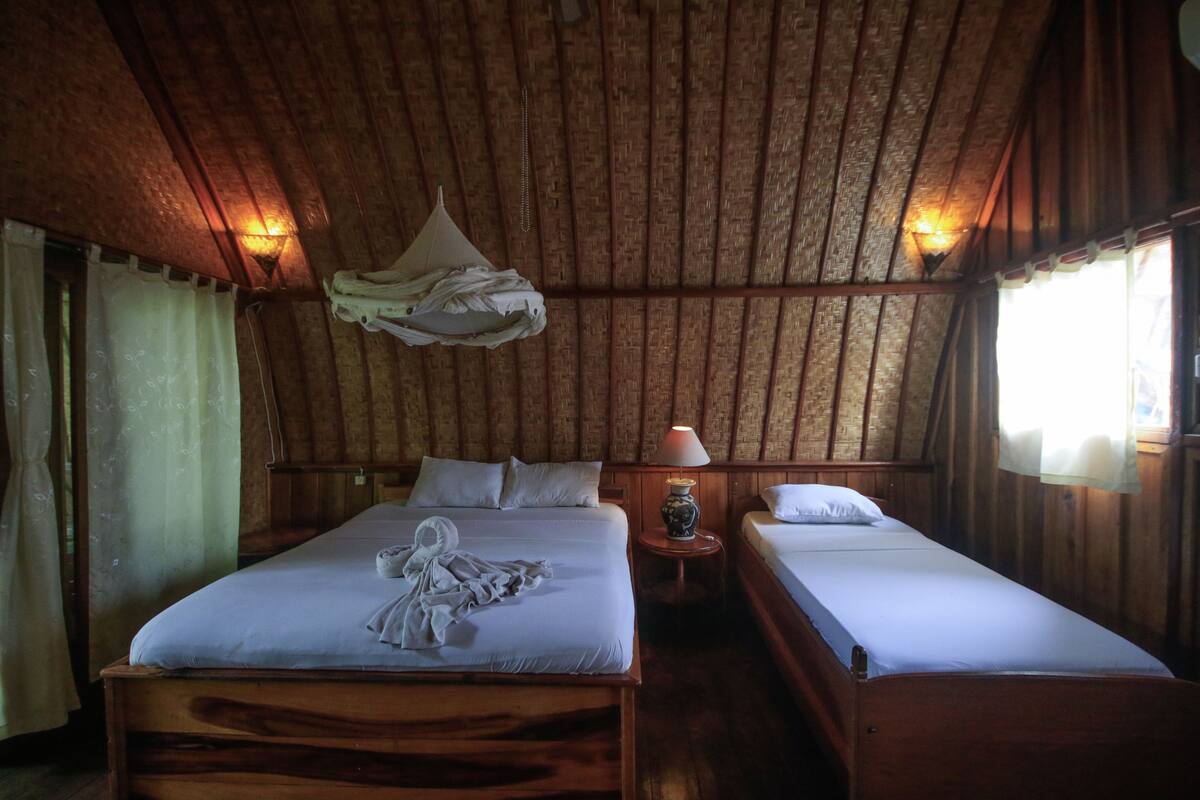 A rustic room with two wooden beds, a hanging mosquito net, and a lamp on a side table.