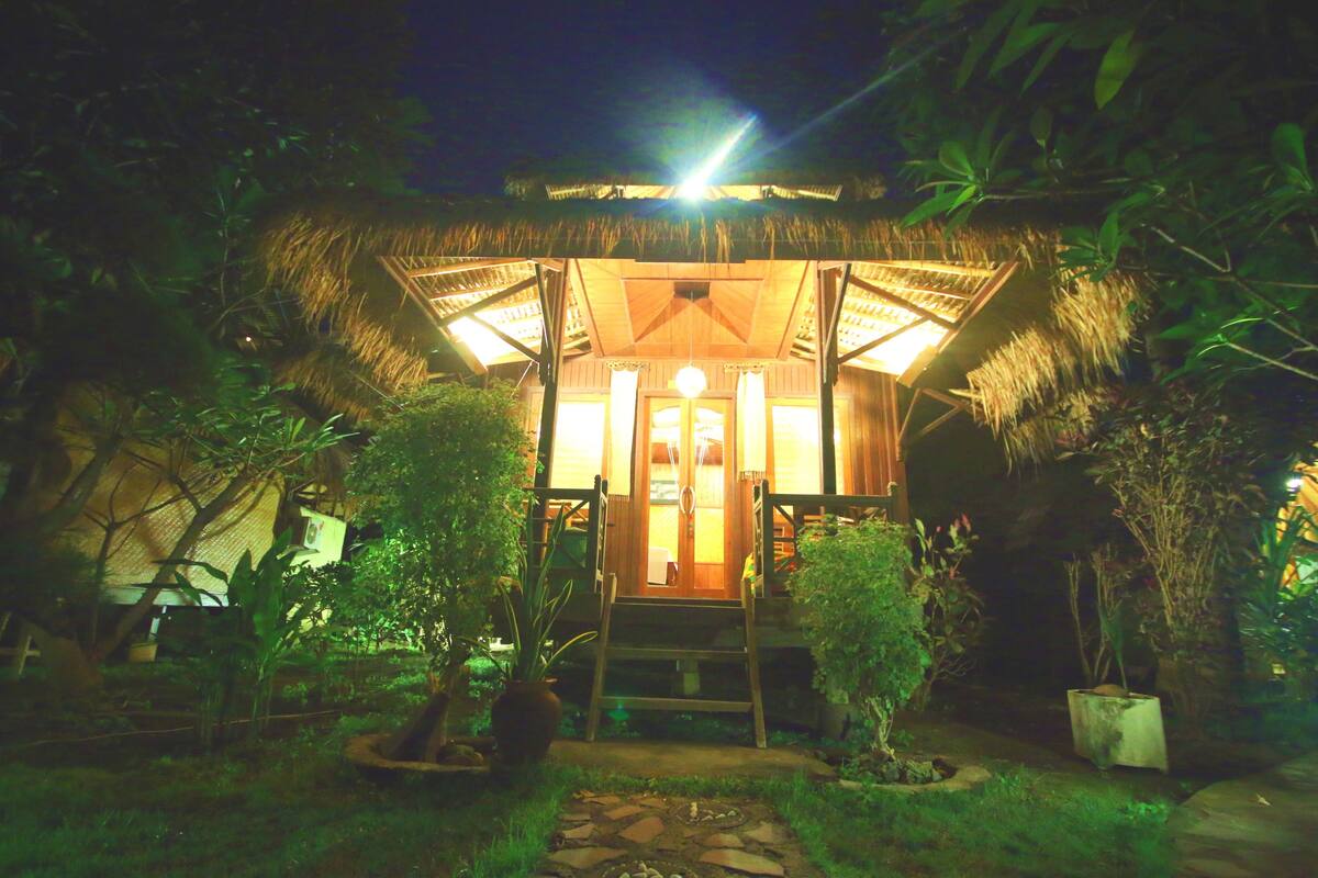 A thatched-roof structure with a staircase leading to a door, surrounded by greenery.