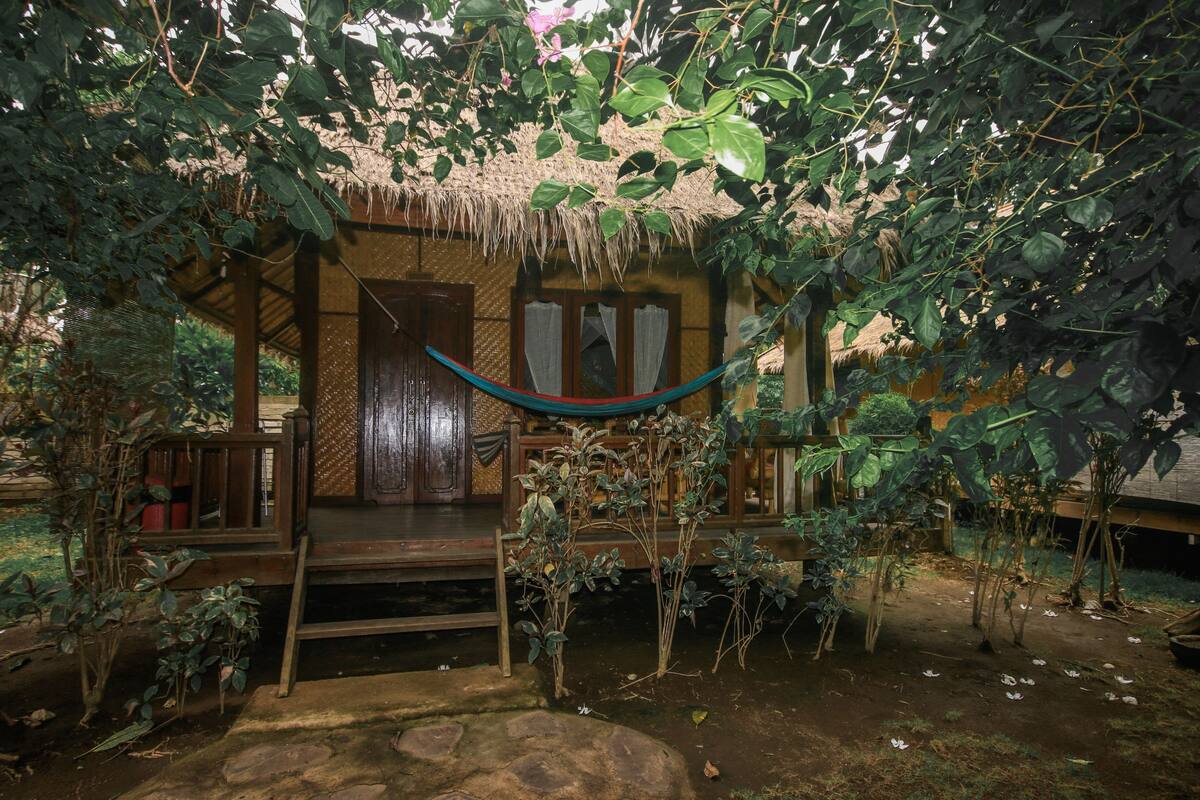 A rustic wooden house with a thatched roof, a wooden porch, and a hammock.