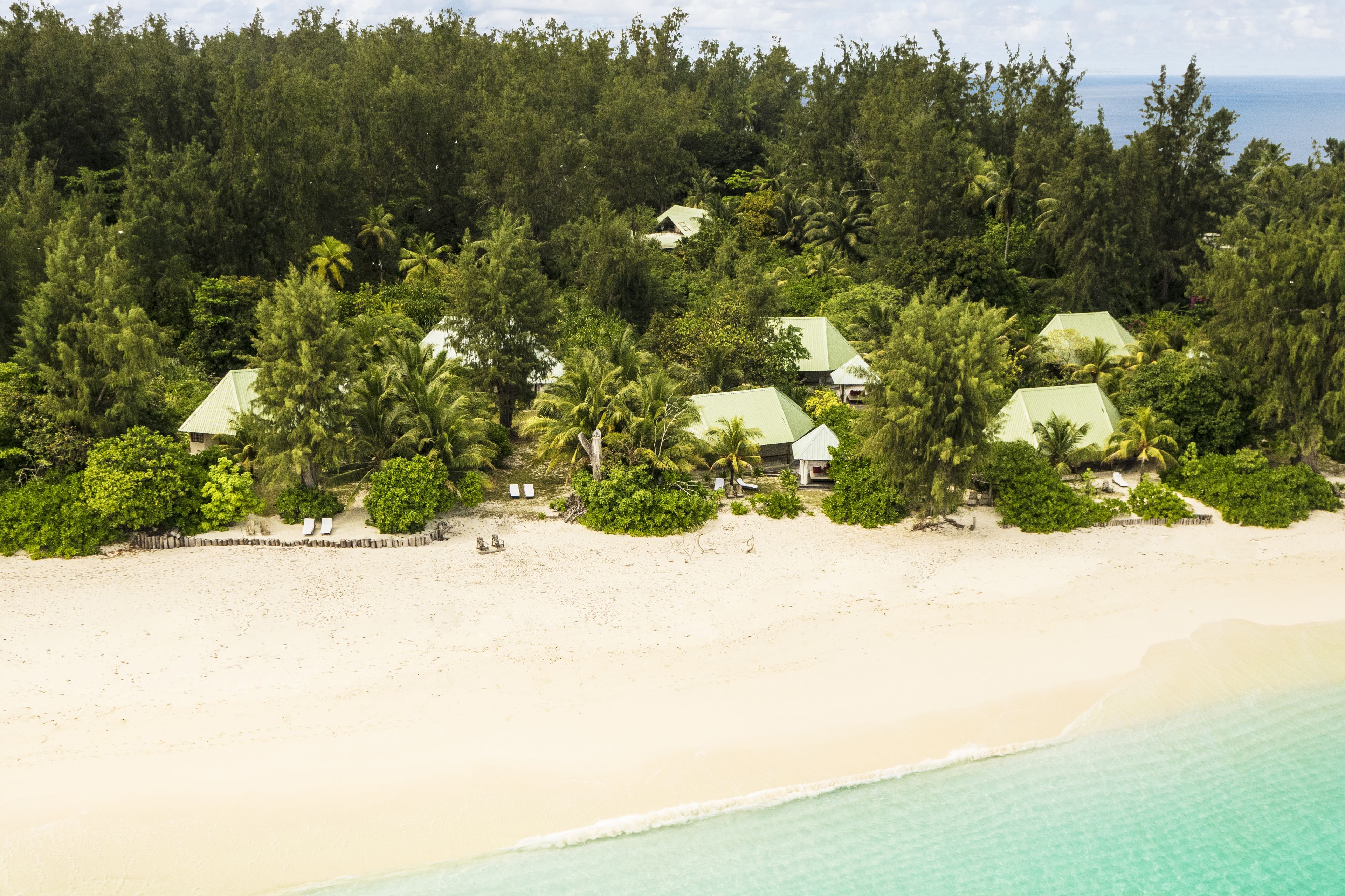 A tropical resort with multiple bungalow-style buildings surrounded by dense greenery and palm trees, overlooking a pristine beach.