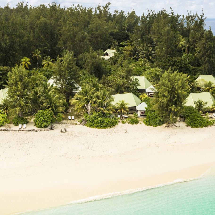 A tropical resort with multiple bungalow-style buildings surrounded by dense greenery and palm trees, overlooking a pristine beach.