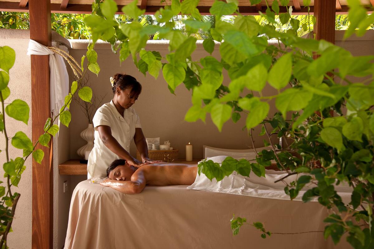 A person receiving a massage in a room with a wooden ceiling, a bed, and a small table with a candle and a basket.