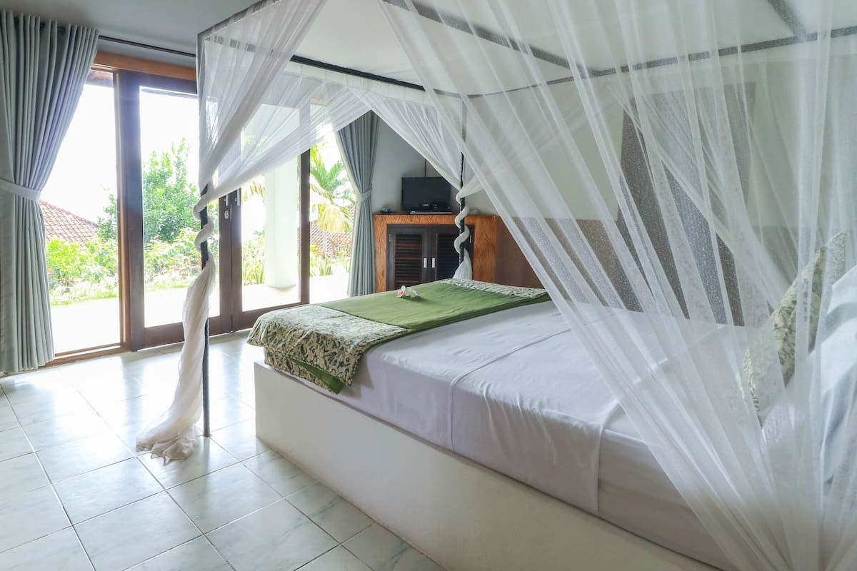 A bedroom with a bed under a canopy, a television on a wooden cabinet, and a view of greenery outside.