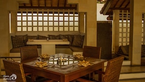 Un comedor con una mesa de madera preparada para cuatro personas, rodeada de sillas de mimbre. Se observa una banca con cojines al fondo, y un ventanal con celosías sobre la mesa.