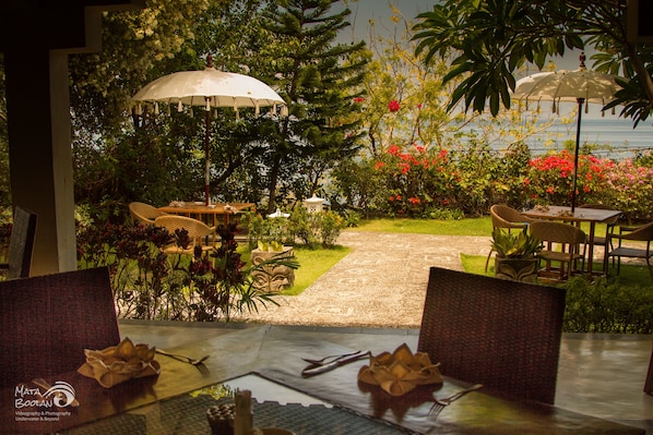 A dining area with tables and chairs, surrounded by lush greenery and a view of the sea.