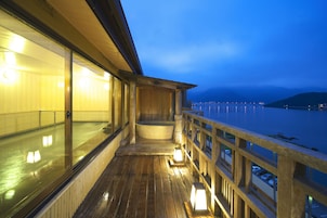 A wooden deck with a glass balustrade overlooking a body of water, a hot tub, and a wooden cabin structure.
