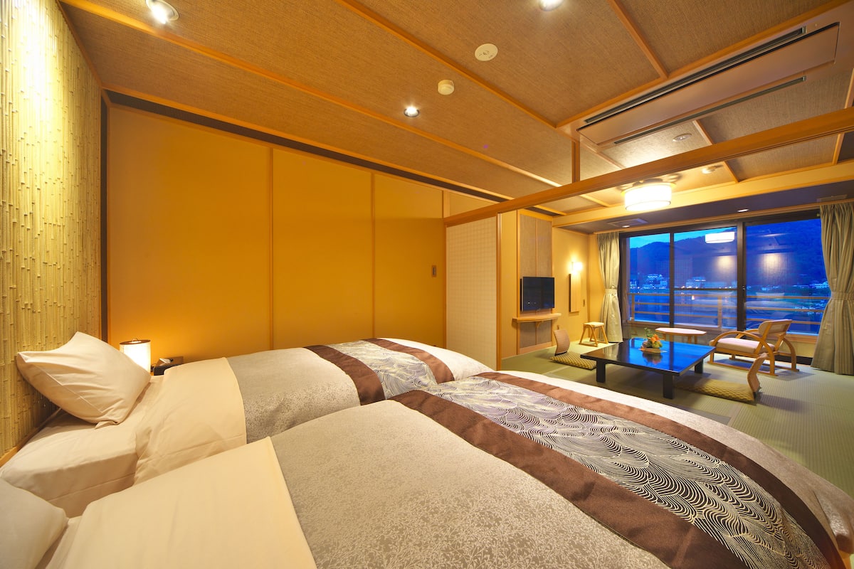 A hotel room with two beds, a small table, a chair, and a large window with a view.