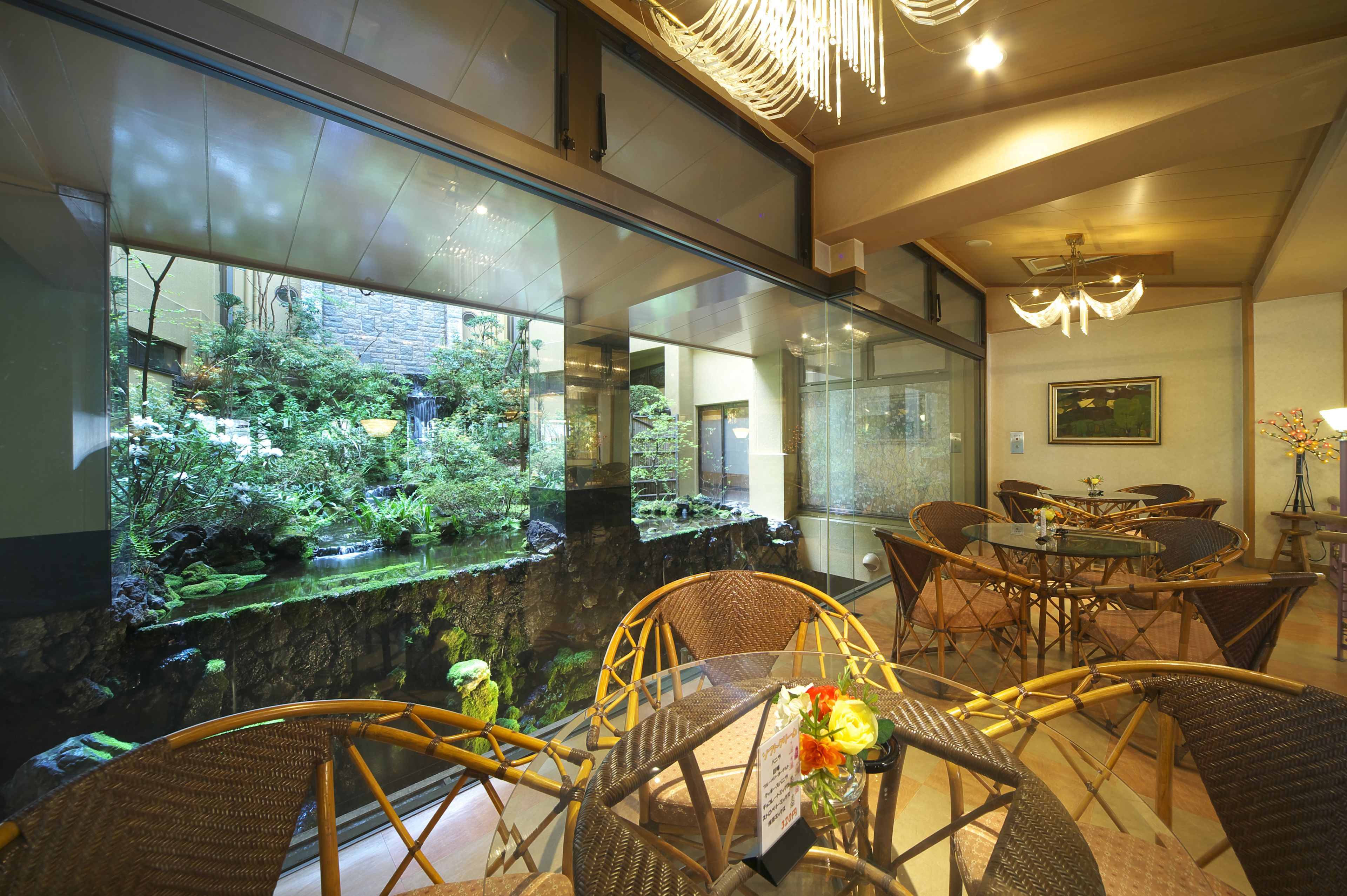 A restaurant with wicker furniture, a glass wall with a view of greenery, and a chandelier.