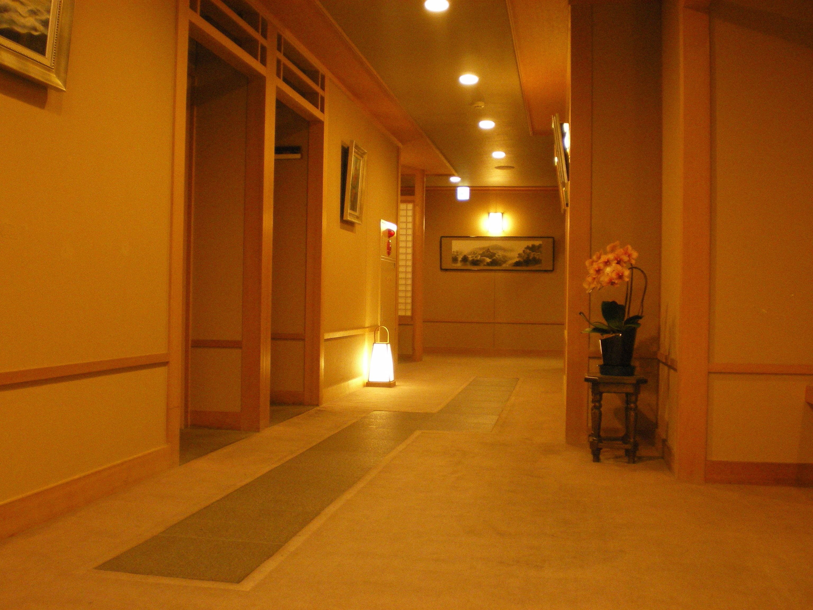 Un pasillo de hotel con piso alfombrado, paneles de madera, una lámpara encendida, un cuadro en la pared y un florero con flores sobre una mesita.
