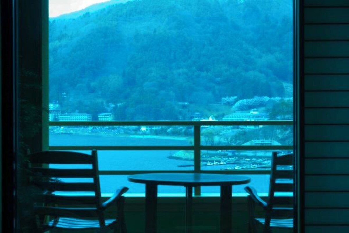 Una habitación con un ventanal amplio que ofrece vistas a un lago y colinas boscosas. En primer plano, se ve una mesa redonda y dos sillas.