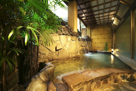 A tranquil indoor hot spring area with a stone waterfall, lush greenery, and a wooden interior.
