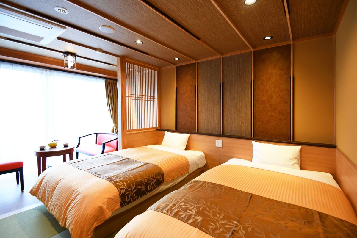 A hotel room with two beds, a small table, a chair, and a view of the outside.