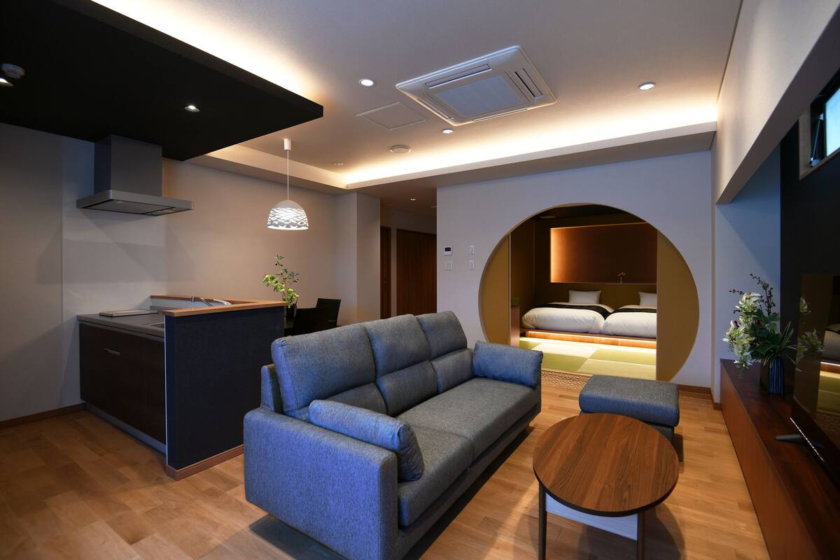 A modern hotel room with a sofa, a small wooden table, a kitchen area, and a circular seating area with a bed.