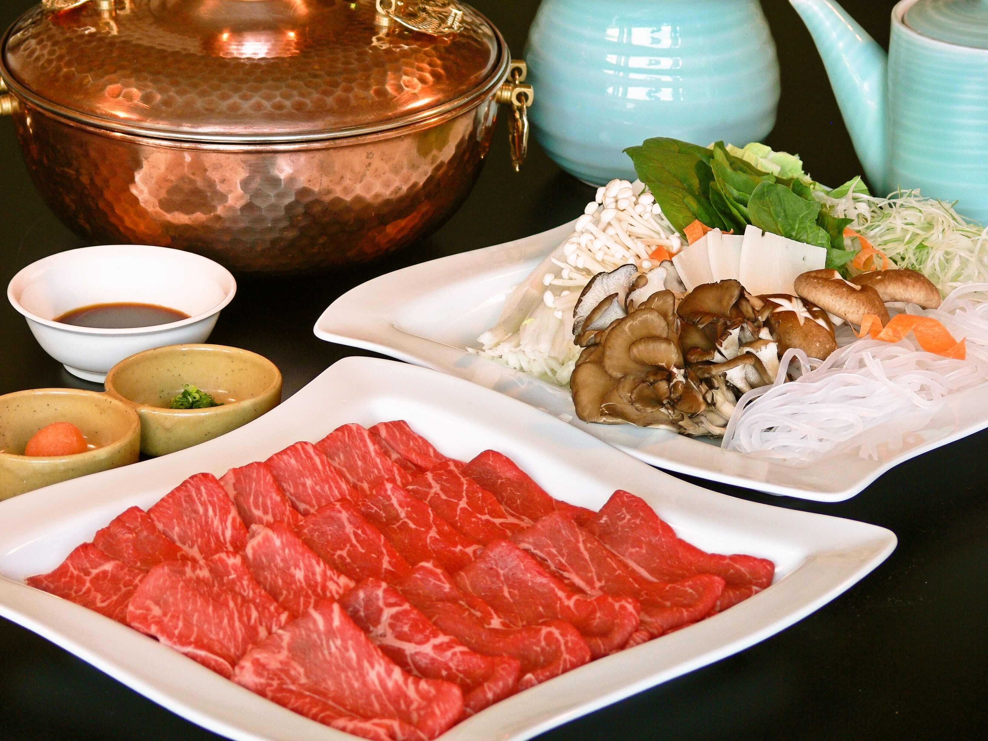 A table with various food items including sliced meat, mushrooms, and vegetables, accompanied by bowls of sauces.