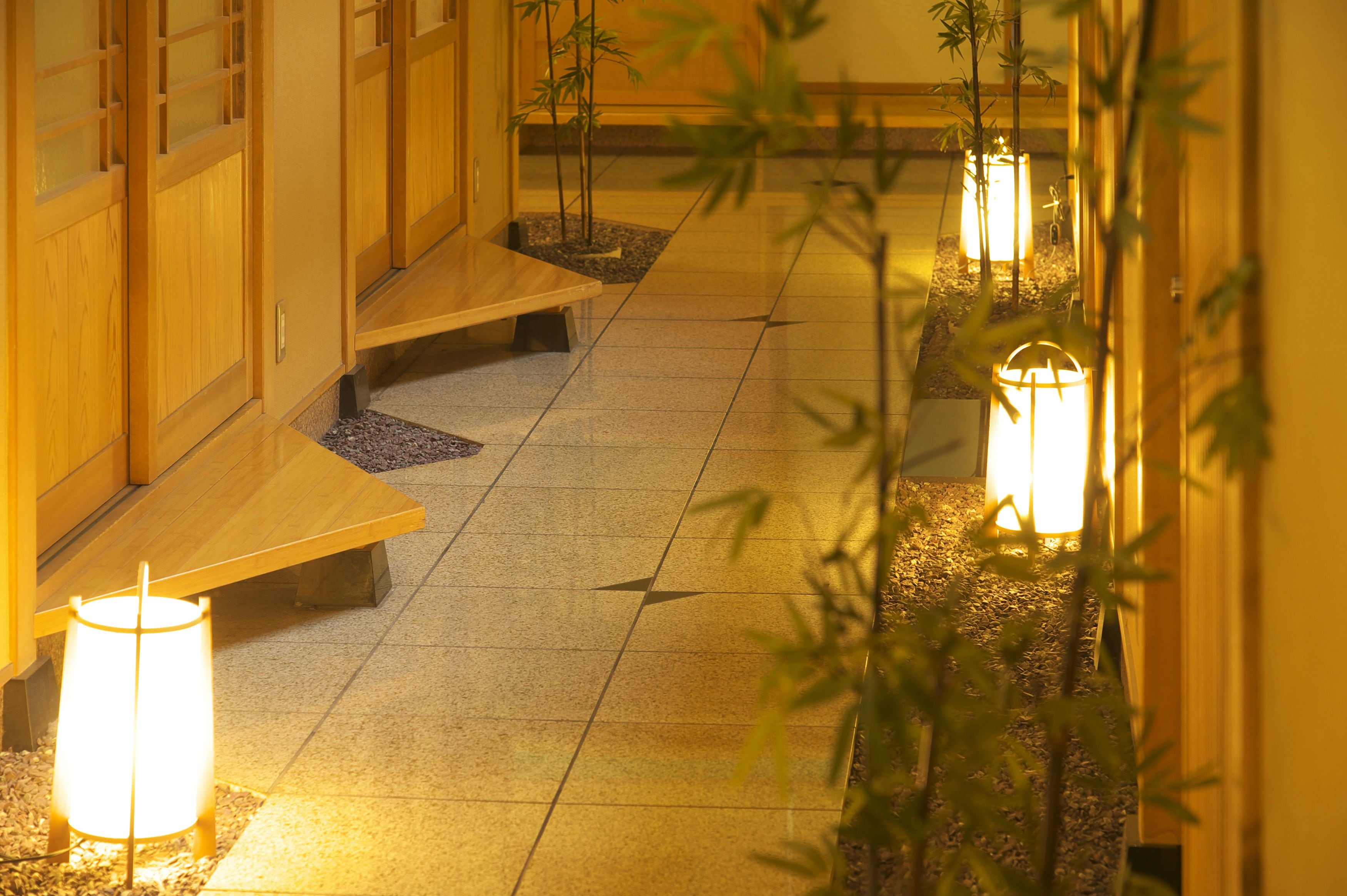 A corridor with wooden benches and illuminated lanterns, leading to a traditional building with sliding doors.