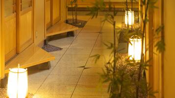 A corridor with wooden benches and illuminated lanterns, leading to a traditional building with sliding doors.
