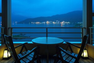 Un balcón con mesa y sillas, con vista a un cuerpo de agua y luces de la ciudad a lo lejos.