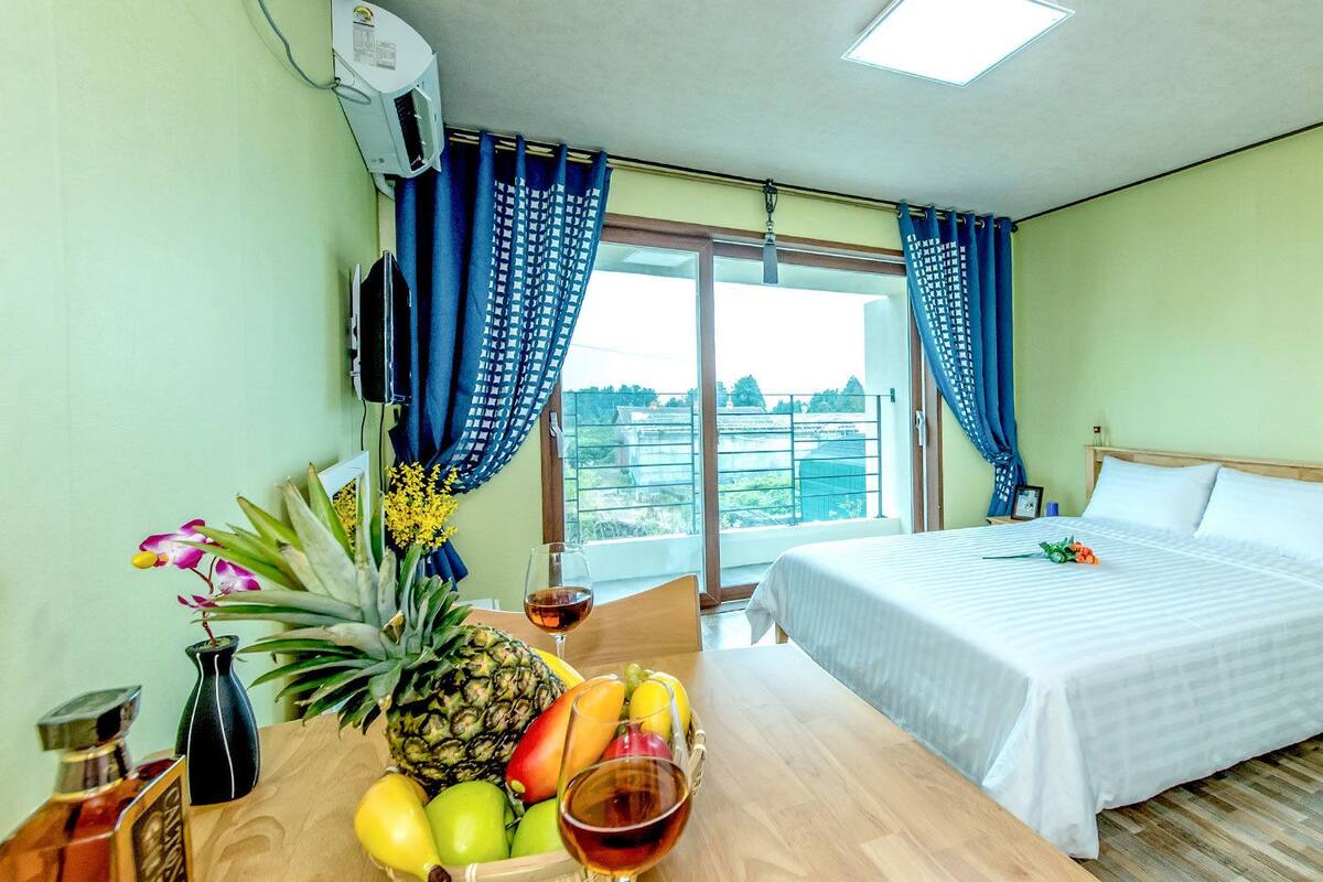 Une chambre d’hôtel avec un lit, une table avec des fruits et une bouteille, et une fenêtre avec des rideaux.