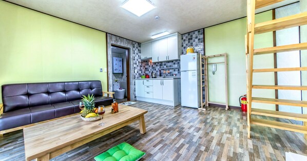 Ein kleiner Wohnraum mit einer Couch, einem Holztisch mit Früchten und Getränken, einer Küchenzeile und einer Holzleiter.
