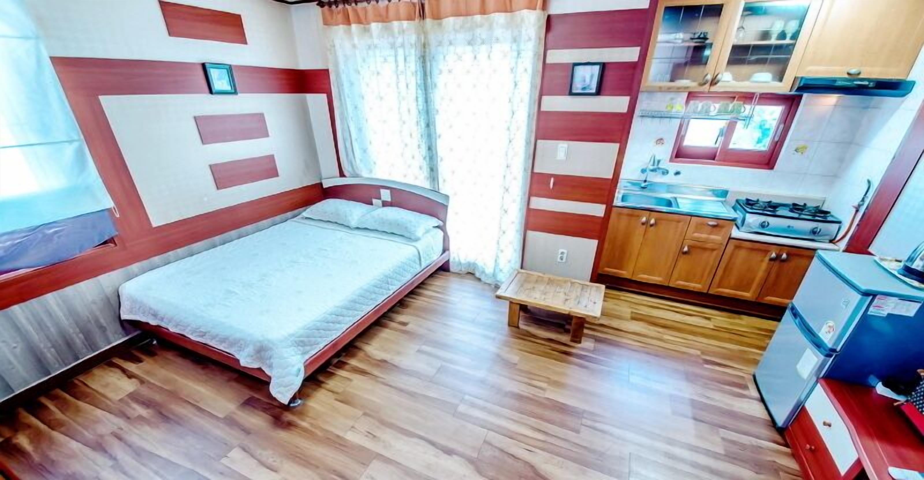 A compact room with a bed, a small bench, a kitchenette with a stove, and wooden cabinets.