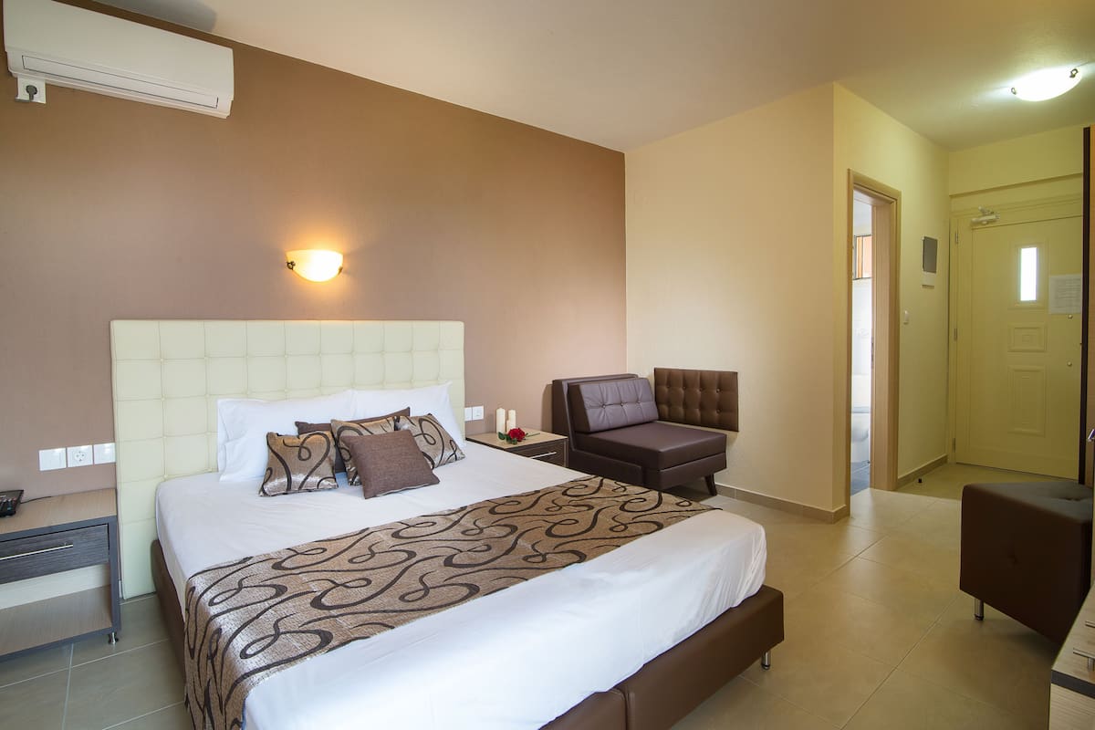 A hotel room with a large bed, a bedside table with a lamp, a chair, and a door leading to another room.