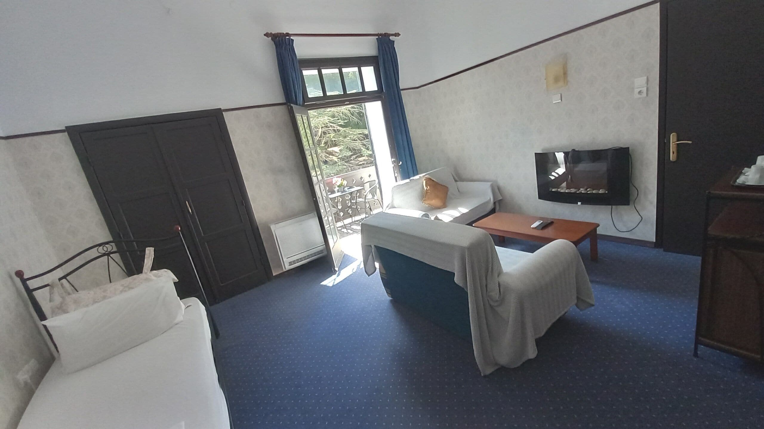 A hotel room with a bed, a small table, a TV, and a balcony with a view of trees.
