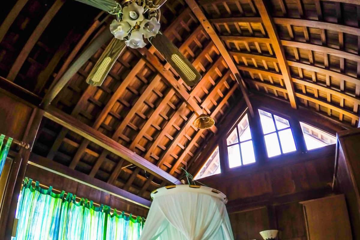 A four-poster bed with a canopy, a wooden ceiling with a crisscross pattern, and a window with curtains.