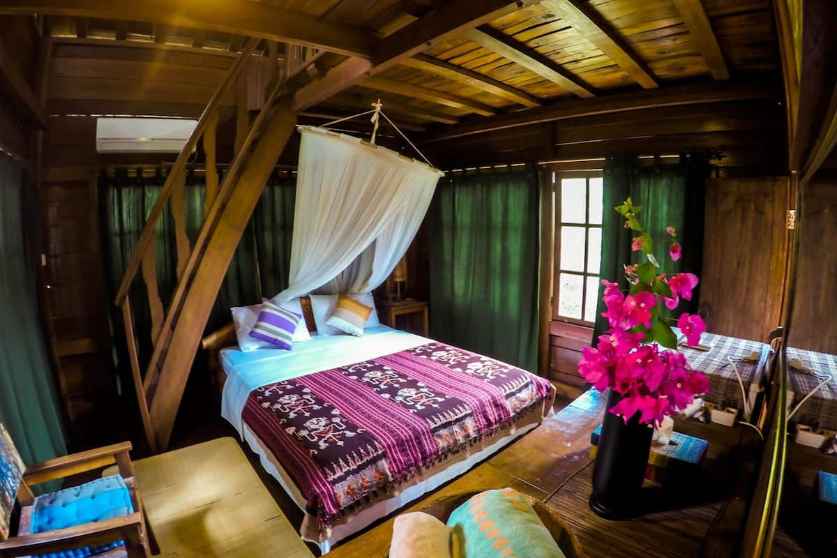A wooden cabin bedroom with a bed, a canopy, a chair, a table, and a potted plant.
