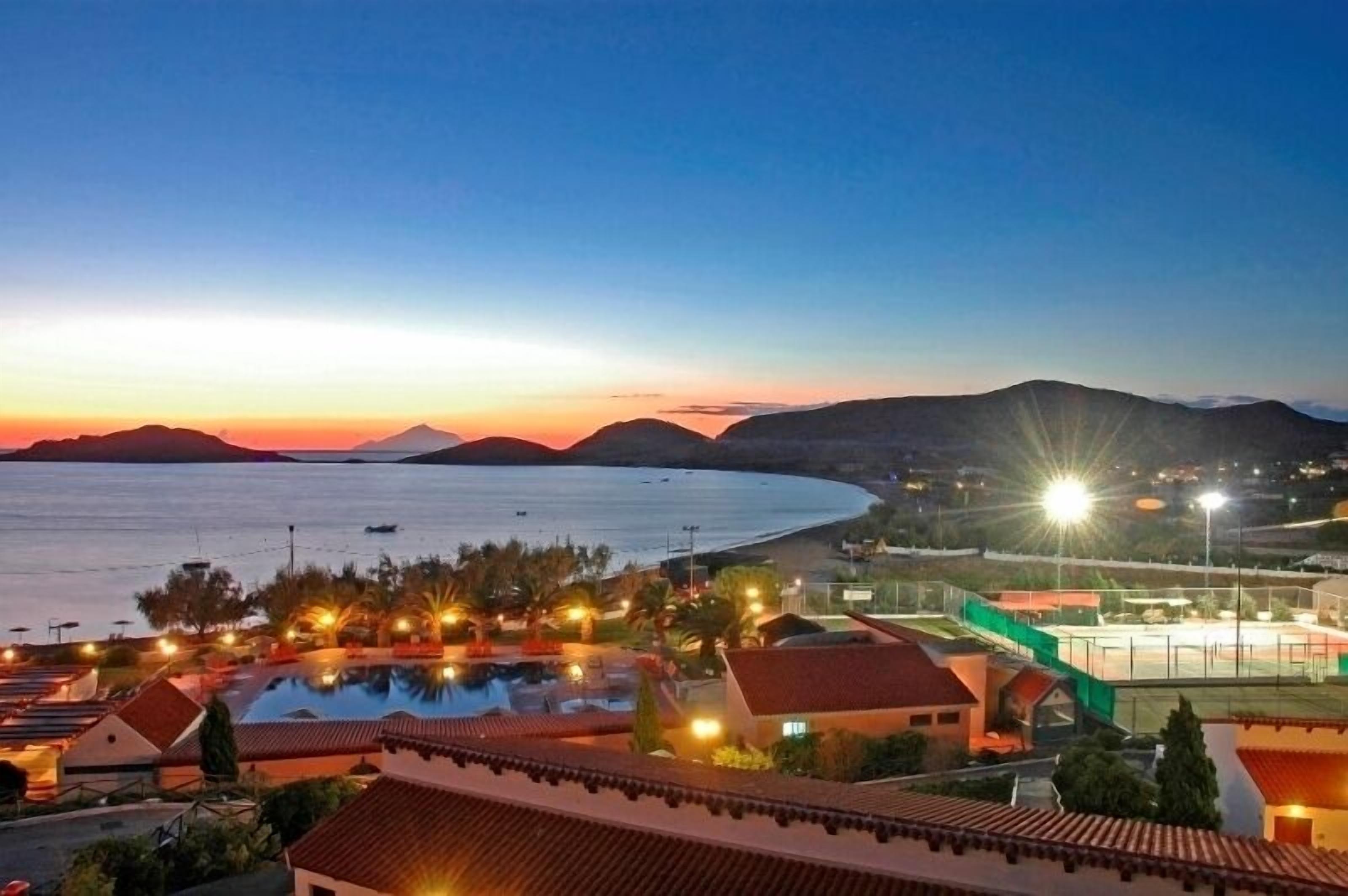 A resort with a large swimming pool, a tennis court, and a view of the sea at sunset.
