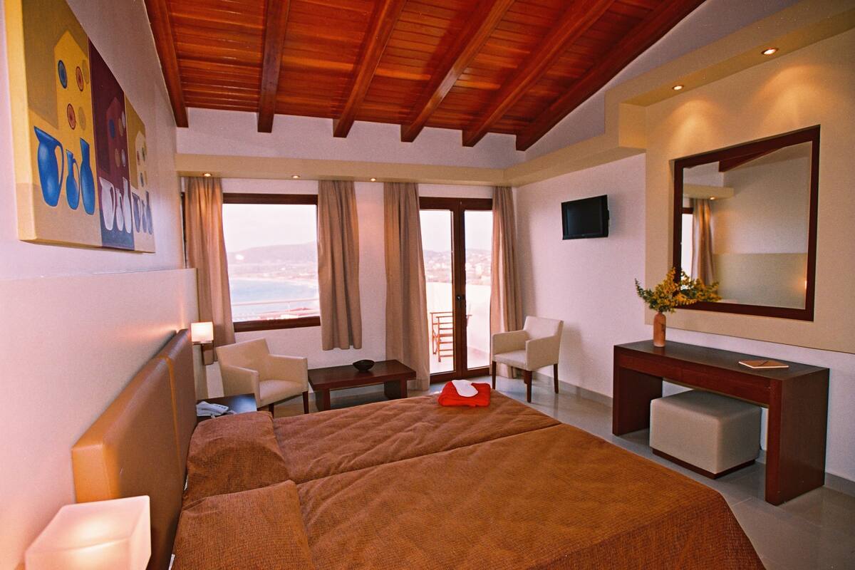 A hotel room with a large bed, a desk, two chairs, a small table, and a view of the sea.