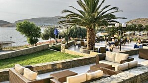 An outdoor seating area with wicker furniture and cushions, a palm tree, and a view of the sea. - Lemnos Village Resort (Lemnos)