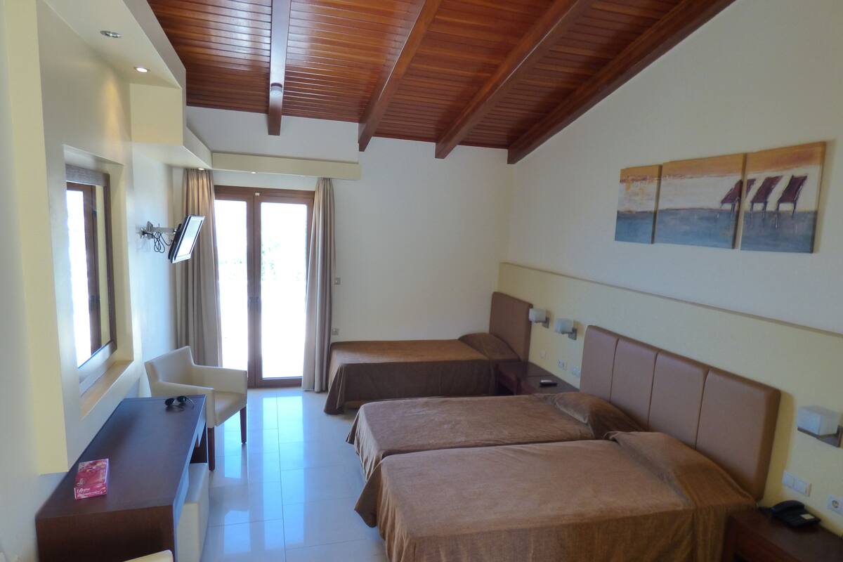 A hotel room with two beds, a desk, and a chair. There is a window with curtains, a ceiling with wooden beams, and a painting on the wall.