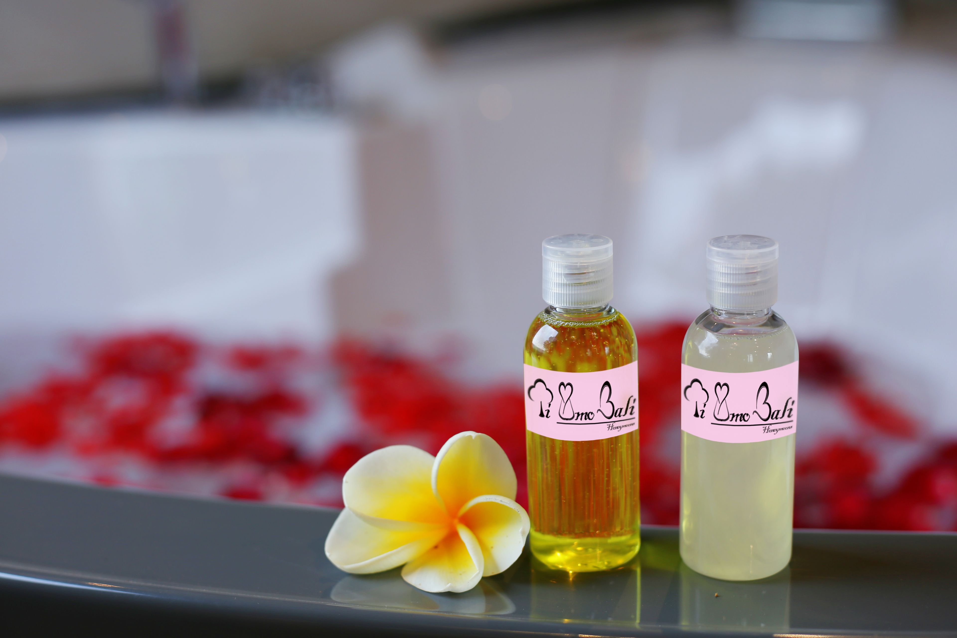 Two bottles of 'Irm Bali' on a reflective surface with a flower and red petals in the background.